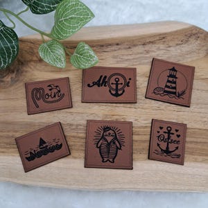 Könnte beinhalten: Sechs braune Lederaufnäher mit maritimen Motiven. Die Aufnäher zeigen Bilder von Anker, Leuchtturm, Segelboot und Seehund. Texte sind "Moin", "Ahoi", "Nordsee" und "Ostsee".
