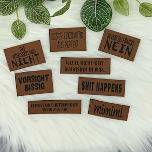 Könnte beinhalten: Acht braune Leder-Patches mit schwarzem Text. Die Patches zeigen verschiedene Sprüche, darunter "Das interessiert mich nicht", "Vorsicht bissig", "Lieber einzigartig als perfekt", "Bevor du fragst: Nein", "Wecke nicht den Wahnsinn in mir...", "Shit happens", "Kannst du subtrahieren? Dann zieh Abi" und "mimimi".