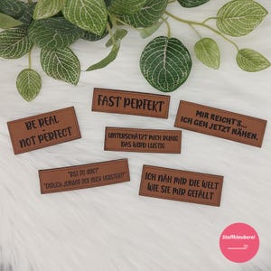 Könnte beinhalten: Sechs braune Lederpatches mit verschiedenen Sprüchen in schwarzer Schrift. Die Sprüche lauten: "Be Real Not Perfect", "Fast Perfekt", "Unterschätzt mich ruhig das wird lustig", "Mir reicht's... Ich geh jetzt nähen.", "'Bist du irre?' 'Endlich jemand der mich versteht!'", und "Ich näh mir die Welt wie sie mir gefällt".