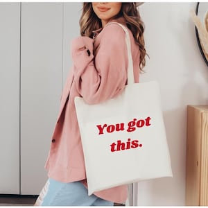 Może przedstawiać: Kremowa torba typu tote z czerwonym napisem "You got this.". Torbę nosi osoba w r&oacute;żowej koszuli. Torba ma prosty design i jest wykonana z jasnej tkaniny.