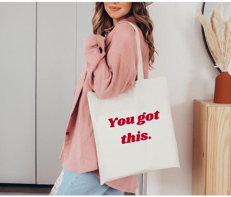 Może przedstawiać: Kremowa torba typu tote z czerwonym napisem "You got this.". Torbę nosi osoba w r&oacute;żowej koszuli. Torba ma prosty design i jest wykonana z jasnej tkaniny.