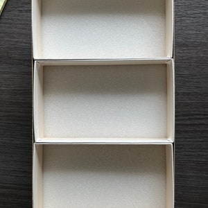 May include: Three-tiered, rectangular storage box with beige cardboard exterior and white foam interior. The box is empty, with each level ready to hold items. The box sits on a dark wood-grain surface.