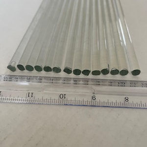 May include: A collection of clear glass straws, each approximately 8 inches long, arranged in a fan shape. A ruler is placed across the bottom of the straws, providing a visual reference for their length. The straws are cylindrical with a smooth, transparent appearance.