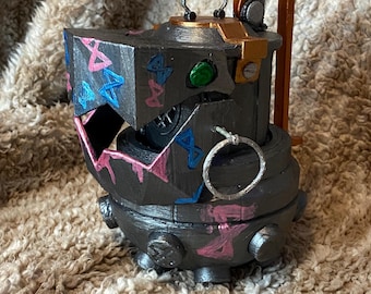 May include: A dark gray, stylized, mechanical object with a geometric design. The object has pink, blue, and gold accents, with a green circular detail. It features a handle and a ring.