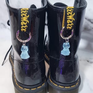 以下が含まれることがあります： 黄色のステッチとプルタブが付いた黒のエナメルブーツのペア。各ブーツには、水色の猫のチャームが付いたシルバーのフープが付いています。ブーツは白とグレーの大理石の表面にあります。