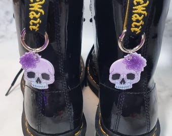 May include: Pair of black leather boots with yellow stitching and a glossy finish. Each boot has a skull charm with a purple flower attached to the pull tab. The charms are on silver hoops.