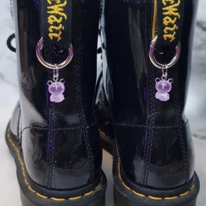 Puede incluir: Par de botas negras de charol con costuras amarillas y una etiqueta amarilla y negra que dice "Dr. Martens". Cada bota tiene un aro plateado con un pequeño dije de oso morado translúcido.