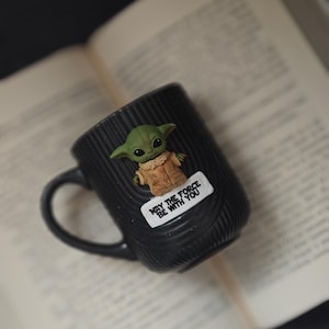 Pu&ograve; includere: Tazza in ceramica nera con superficie strutturata, raffigurante un personaggio verde Grogu e la scritta "MAY THE FORCE BE WITH YOU". La tazza &egrave; appoggiata su un libro aperto.