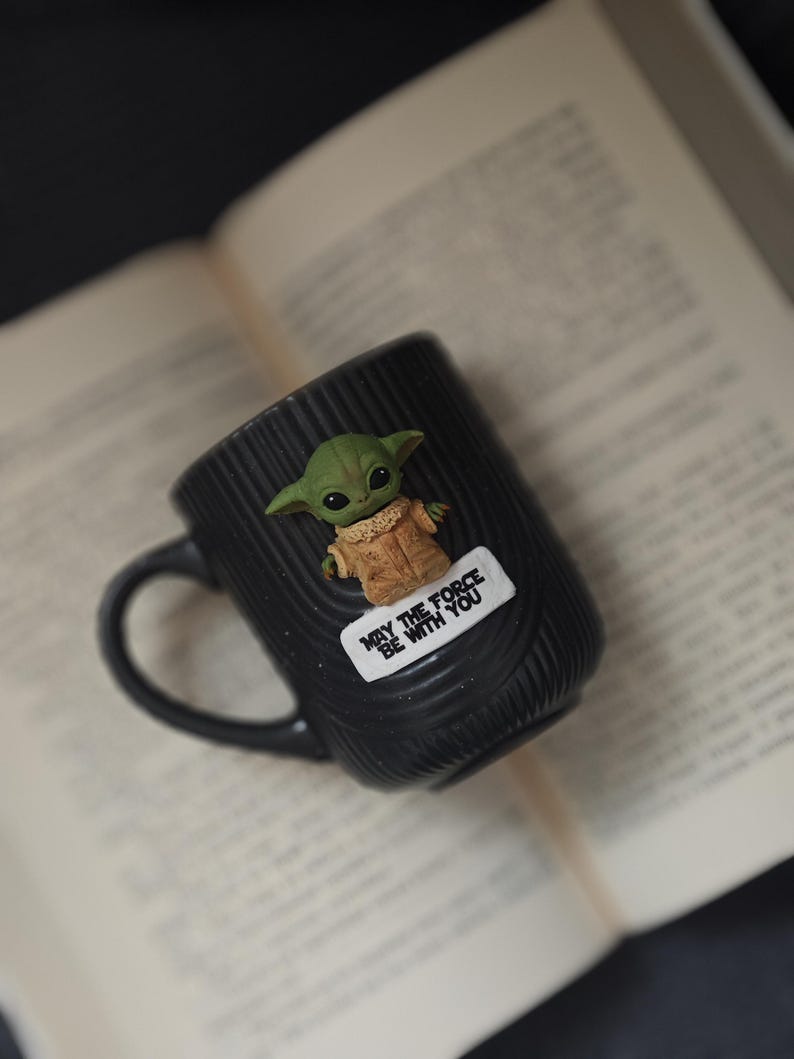 Pu&ograve; includere: Tazza in ceramica nera con superficie strutturata, raffigurante un personaggio verde Grogu e la scritta "MAY THE FORCE BE WITH YOU". La tazza &egrave; appoggiata su un libro aperto.