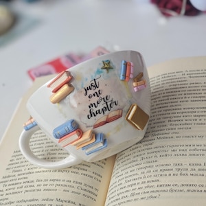 Può includere: Tazza in ceramica bianca decorata con libri in miniatura di vari colori e una stella dorata. La tazza presenta la scritta "just one more chapter" in nero. La tazza è appoggiata su un libro aperto.