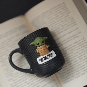 Pu&ograve; includere: Tazza in ceramica nera con un personaggio alieno verde e la scritta "MAY THE FORCE BE WITH YOU". La tazza ha un manico ed &egrave; appoggiata su un libro aperto. Il personaggio alieno ha grandi orecchie e un cappotto beige.