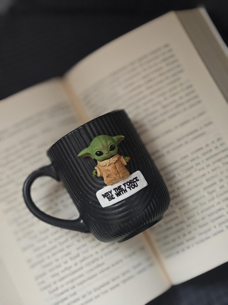 Pu&ograve; includere: Tazza in ceramica nera con un personaggio alieno verde e la scritta "MAY THE FORCE BE WITH YOU". La tazza ha un manico ed &egrave; appoggiata su un libro aperto. Il personaggio alieno ha grandi orecchie e un cappotto beige.