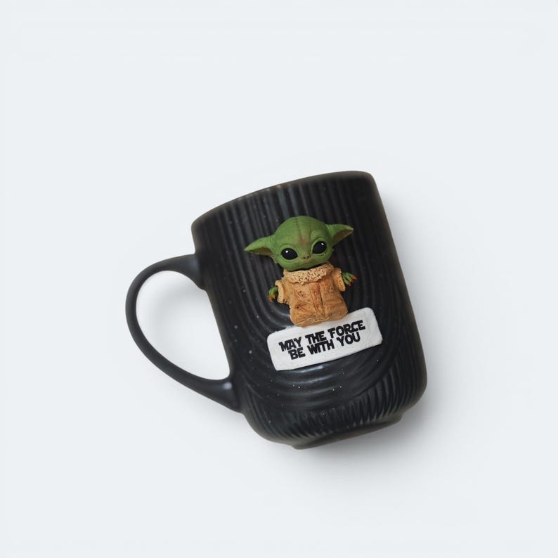 Pu&ograve; includere: Tazza in ceramica nera con una figura di Grogu e la scritta "MAY THE FORCE BE WITH YOU". La tazza ha una superficie strutturata e un manico curvo. La figura di Grogu &egrave; verde con grandi occhi e indossa un cappotto marrone.