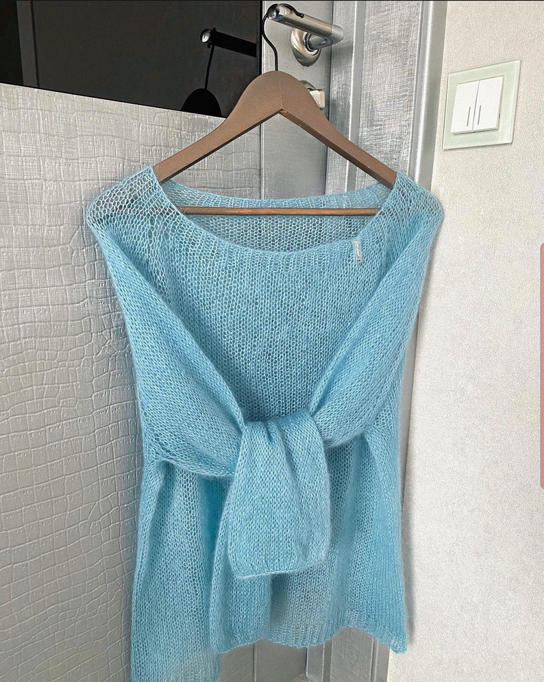 May include: A light blue knitted sweater with a loose fit and a wide neckline. The sweater is made of a soft, delicate yarn and has a slightly textured appearance.