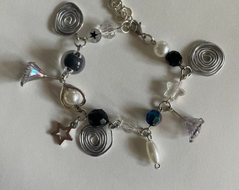 May include: A silver charm bracelet with a variety of charms, including stars, swirls, and beads. The bracelet is made of silver chain and has a lobster clasp closure.