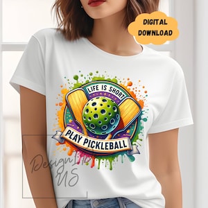 Op de afbeelding: Wit T-shirt met een kleurrijk grafisch ontwerp met twee pickleball-rackets en een bal met de tekst "Life is short, Play Pickleball".