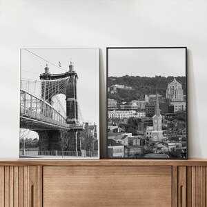 May include: Two black and white photographs in frames. The left photo shows a suspension bridge with a cityscape in the background. The right photo shows a cityscape with a church steeple in the foreground.