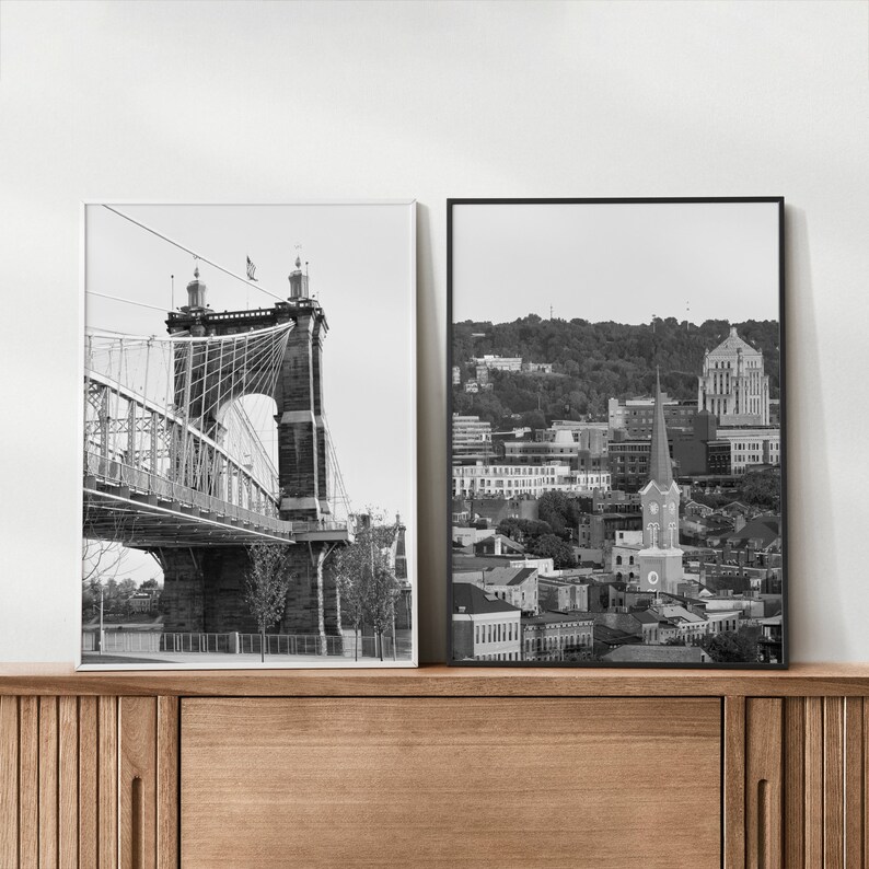 May include: Two black and white photographs in frames. The left photo shows a suspension bridge with a cityscape in the background. The right photo shows a cityscape with a church steeple in the foreground.