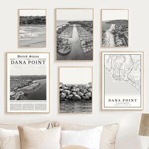 May include: A collection of six framed black and white prints. The prints depict coastal scenes, including a marina, a beach, and a map of Dana Point, California. The prints are displayed on a white wall.