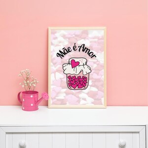 Op de afbeelding: Een ingelijst print met een roze achtergrond en witte hartjes. De print toont een pot gevuld met roze hartjes en de tekst "Mãe é Amor" in zwart.