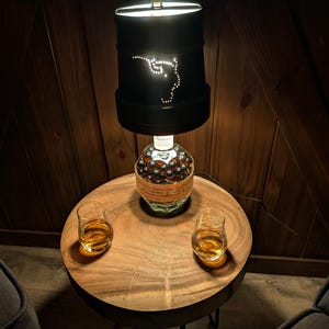 May include: A wooden table with two glasses of amber liquid and a lamp made from a liquor bottle with a black metal shade. The shade has a cut-out design of a star.