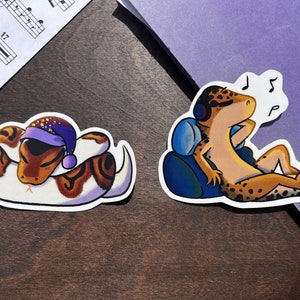 May include: Two stickers featuring cartoon animals. The first sticker shows a brown and white snake curled up asleep wearing a purple and white hat. The second sticker shows a yellow and brown gecko wearing headphones and listening to music. The gecko is sitting on a blue chair with musical notes above its head.