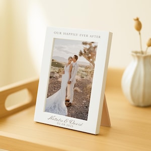May include: A white wooden photo frame with a wedding photograph. The frame is inscribed with "OUR HAPPILY EVER AFTER" at the top and the names "Natalie & David" with the date "02.08.2025" at the bottom. The photo features a newly married couple.