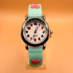 May include: A quartz wristwatch with a mint green silicone strap featuring raised pink heart accents. The round silver-tone case frames a white dial with multicoloured Arabic numerals, black minute markers, and three hands, including a blue second hand.