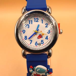 May include: A blue wristwatch with a silver-tone metal case and a white dial featuring blue Arabic numerals. The watch face displays a small rocket graphic and yellow and blue hands. The blue silicone strap features a 3D astronaut and rocket.