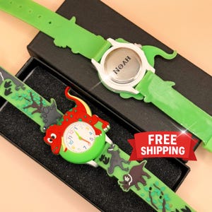 May include: Two children's watches with green straps and dinosaur designs. One watch features a red and green dinosaur on the face, while the other has the name "NOAH" on the back. The straps have dinosaur and tree graphics.