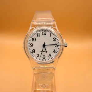 Puede incluir: Un reloj de cuarzo transparente con esfera blanca y números negros. El reloj tiene una correa y una caja de plástico transparente. La esfera muestra los números del 1 al 12, con manecillas negras que indican la hora. La palabra "QUARTZ" está impresa en la esfera.