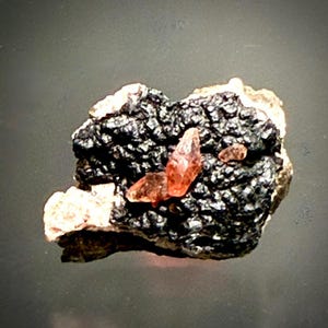 Puede incluir: Un espécimen mineral que presenta un grupo de cristales translúcidos de color naranja rojizo incrustados en una matriz oscura, texturizada y de aspecto metálico. La base muestra tonos tierra claros que contrastan con la superficie oscura y rugosa.