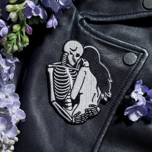 May include: A black leather jacket featuring a white embroidered patch of a skeleton embracing a woman. The jacket is adorned with a bunch of purple lilac flowers.