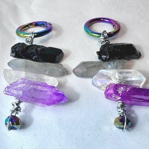 May include: Two rainbow iridescent metal rings with black, white, and purple crystal charms hanging from them. The charms are attached with silver wire and beads.