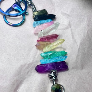 May include: A keychain featuring a collection of colorful crystal points. The crystals are arranged in a vertical line, showcasing a spectrum of colors including purple, blue, green, yellow, pink, and clear. The keychain has a metallic iridescent clasp and a silver wire wrap.