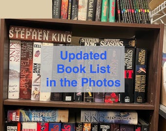 May include: A dark brown wooden bookshelf filled with numerous Stephen King books arranged in rows. The image features text overlays stating Updated Book List in the Photos and Book List, Prices & Conditions with a black arrow pointing right.