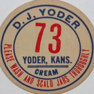 May include: A round, vintage cream-colored label with blue and red text. The label reads "D. J. Yoder" at the top, with the number "73" in the center. The text "Yoder, Kans. Cream" is also present.