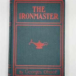 May include: A hardcover book titled "The Ironmaster" with red lettering and a red border on a teal cover. The cover features a red genie lamp illustration and the author's name, Georges Ohnet.