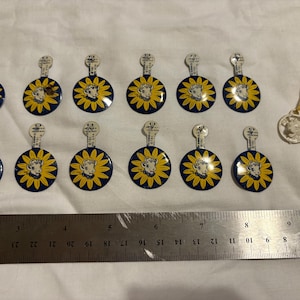 May include: A collection of vintage circular pins with a blue and yellow sunflower design and a cartoon character in the center. A metal ruler is placed below the pins. One pin is attached to a string.