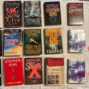 May include: A collection of Stephen King books, including titles like "Mr. Mercedes," "Doctor Sleep," and "Pet Sematary." The books are hardcover and paperback, with various cover designs and colors. The books are arranged on a white surface.