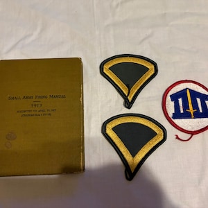 May include: A vintage Small Arms Firing Manual from 1913, alongside two black and gold chevrons and a circular patch with a sword and building design. The manual is a faded olive green color.