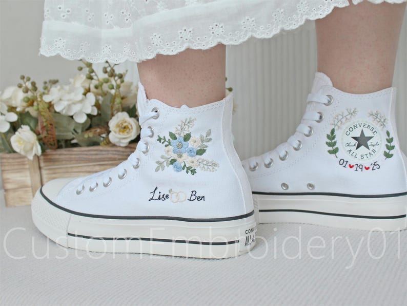 Może przedstawiać: Białe, wysokie trampki Converse z haftowanymi wzorami kwiatowymi i imionami "Lise & Ben". Jedno z but&oacute;w ma haftowane logo Converse i datę "01.19.15".