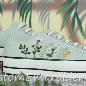 Peut inclure: Une paire de baskets en toile vert clair avec des semelles et des lacets blancs. Les baskets sont décorées de fleurs et de feuilles brodées dans des tons de vert, blanc et jaune.