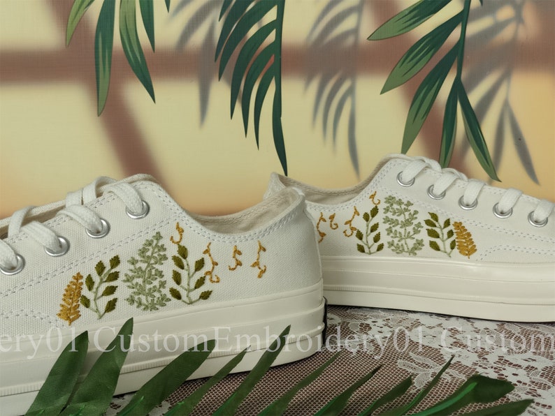 May include: White canvas sneakers with green and yellow embroidered floral designs. The shoes have white laces and a white rubber sole.