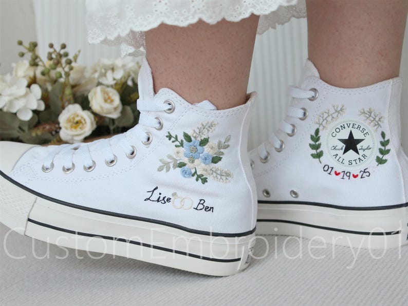 Może przedstawiać: Białe, wysokie trampki Converse z haftowanymi wzorami kwiatowymi i tekstem "Lise & Ben" na jednym bucie i "Converse All Star 01 &hearts; 19 &hearts; 25" na drugim.