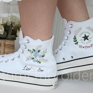 Może przedstawiać: Białe, wysokie trampki Converse z haftowanymi wzorami kwiatowymi i imionami "Lisa" i "Ben". Jedno z but&oacute;w ma haftowane logo Converse i datę "01&hearts;19&hearts;25".