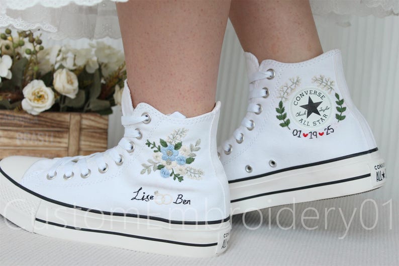 Może przedstawiać: Białe, wysokie trampki Converse z haftowanymi wzorami kwiatowymi i imionami "Lisa" i "Ben". Jedno z but&oacute;w ma haftowane logo Converse i datę "01&hearts;19&hearts;25".