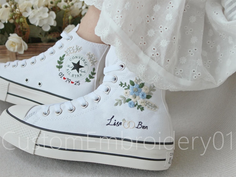 Może przedstawiać: Białe, wysokie trampki Converse z haftowanymi wzorami kwiatowymi i tekstem "Lise & Ben" oraz "01&hearts;19&hearts;25".