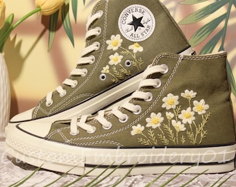 May include: Olive green high-top sneakers with white laces and soles. The sneakers feature embroidered white and yellow daisy flowers. The Converse All Star logo is visible on the side of the shoe.