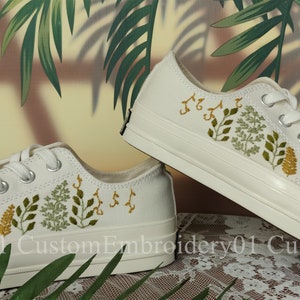 May include: A pair of white canvas sneakers with embroidered green and yellow floral designs. The shoes are a classic style with laces and a white rubber sole.